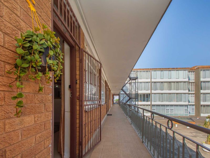 A covered exterior balcony walkway featuring brick wall finishes, metal security gates, and a metal railing overlooking the apartment complex courtyard.