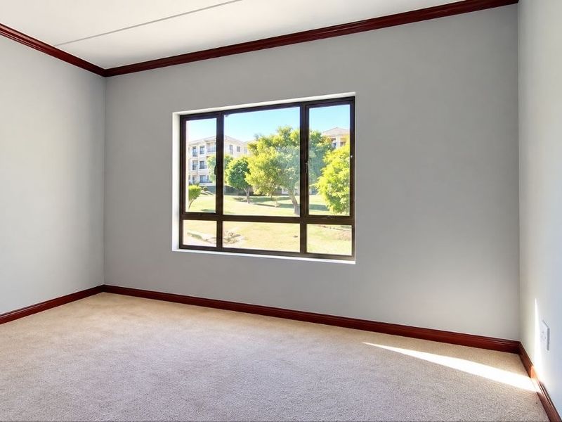 A bright, empty bedroom featuring neutral walls, carpeted flooring, dark timber trim, and a large window with views of the surrounding complex grounds.