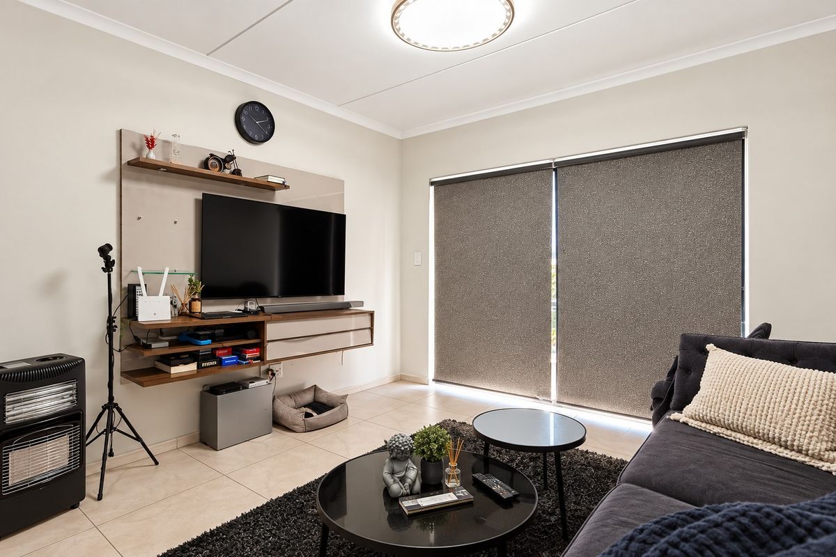 A well-maintained living room featuring tiled flooring, a modern built-in media console, and large windows fitted with roller blinds.