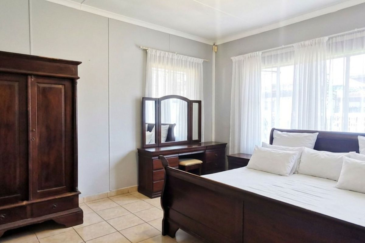 A spacious bedroom featuring neutral wall finishes, tiled flooring, and large windows with curtain rails, offering a bright and functional living space.