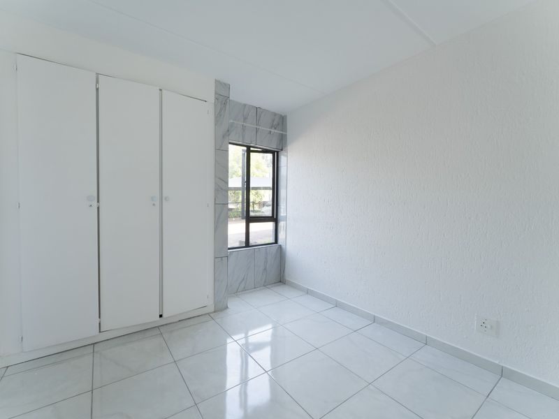 A bright, empty bedroom featuring white built-in cupboards, modern white tiled flooring, and a window framed with marble-look tiles.