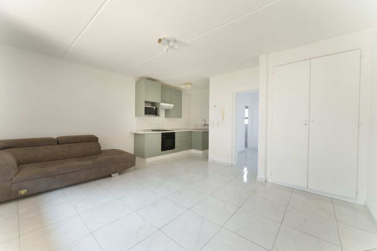 Open plan living area featuring a compact kitchen with built-in cabinetry, integrated appliances, and light-colored tiled flooring throughout.
