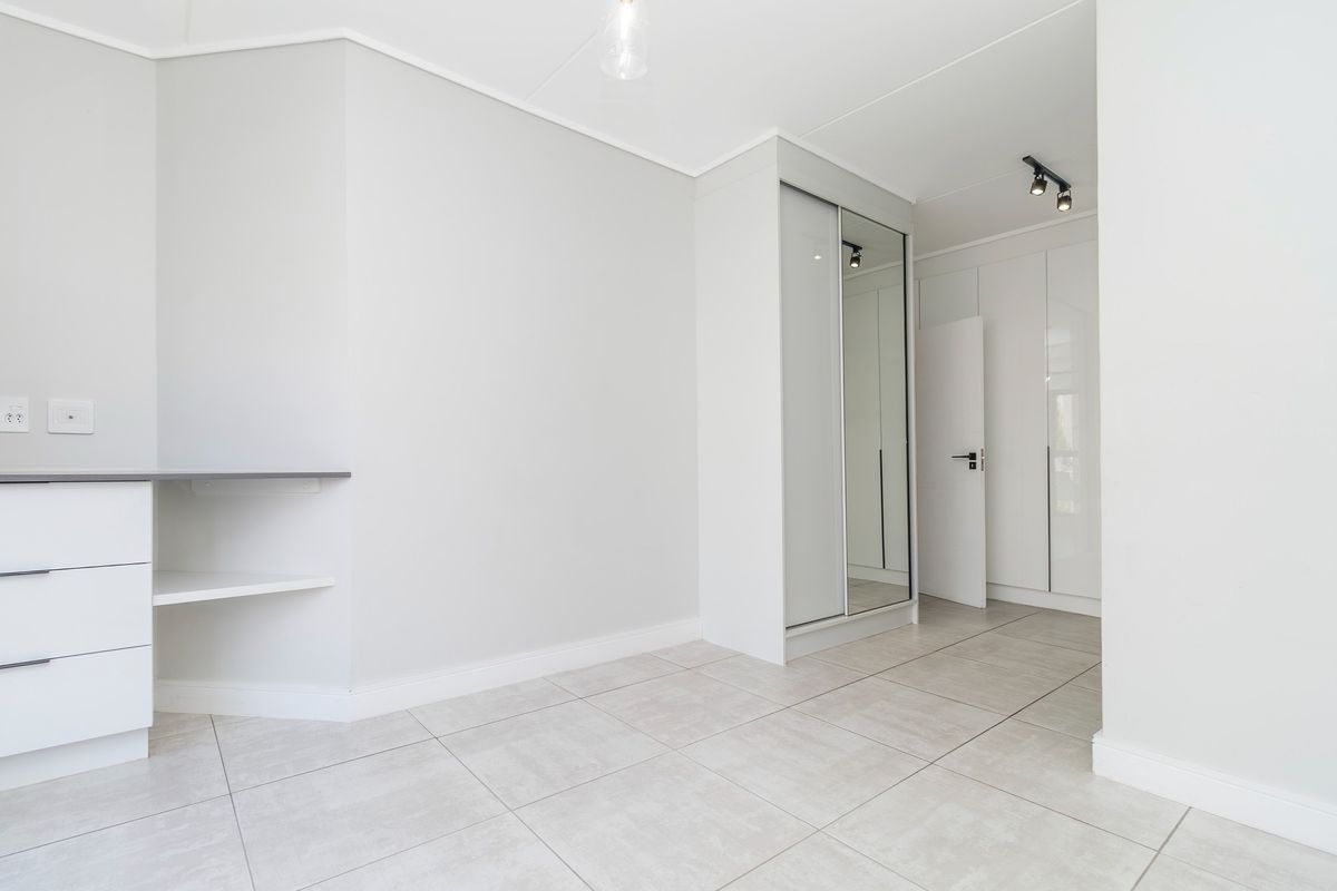 A bright, modern bedroom featuring neutral tiled flooring, ample built-in cupboard space with mirrored doors, and a convenient built-in desk area.