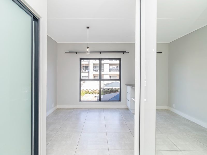 A bright, modern open-plan living area featuring neutral tiled flooring, large aluminium-framed windows, and minimalist built-in cabinetry, offering a clean and versatile space.