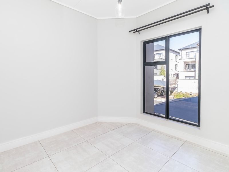 A bright, empty bedroom featuring neutral tiled flooring, a modern black aluminium window frame, and a simple pendant light fixture.