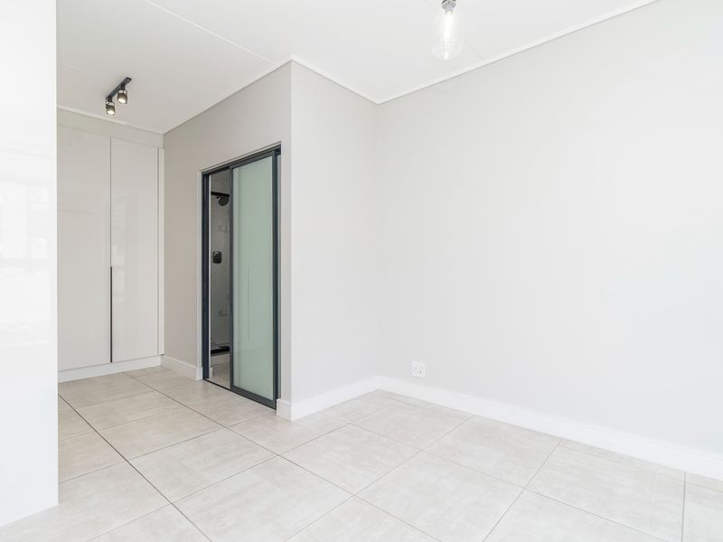 A bright, modern bedroom featuring neutral tiled flooring, built-in cupboards, and a sleek sliding door leading to an en-suite bathroom.
