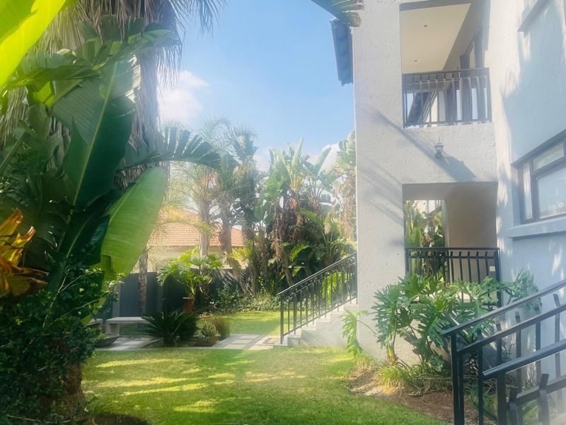 A well-maintained rear garden featuring a lush lawn, mature tropical plants, a paved walkway, and a multi-level balcony with metal railings.