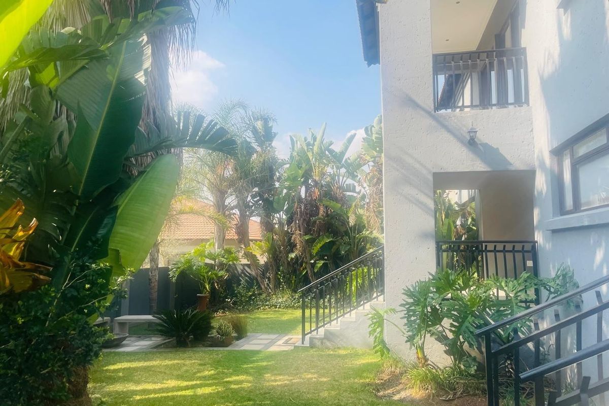 A well-maintained rear garden featuring a lush lawn, mature tropical plants, a paved walkway, and a multi-level balcony with metal railings.