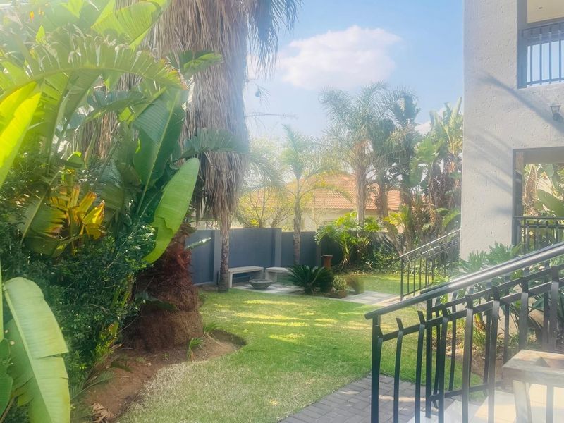 A well-maintained garden area featuring a paved walkway, lush landscaping, and a built-in stone bench, viewed from an elevated staircase.
