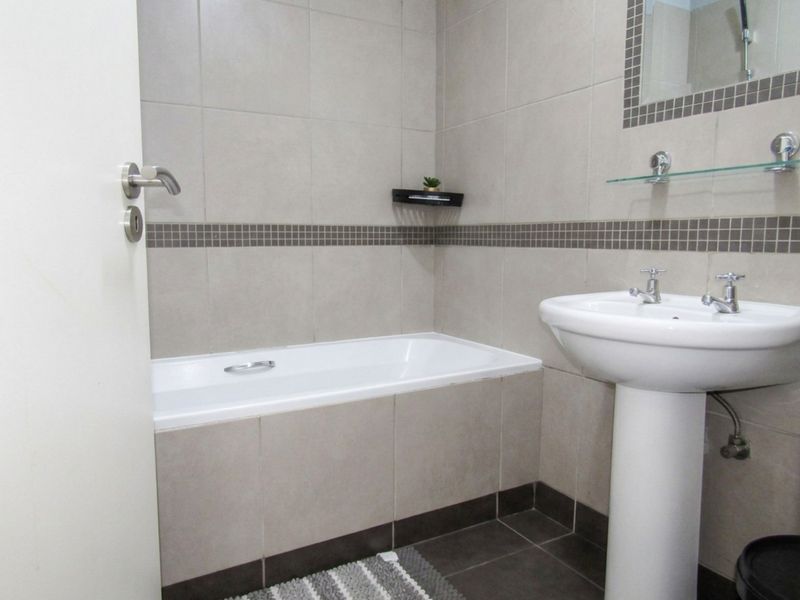 A clean, neutral-toned bathroom featuring a built-in bathtub, pedestal basin, and tiled finishes throughout.