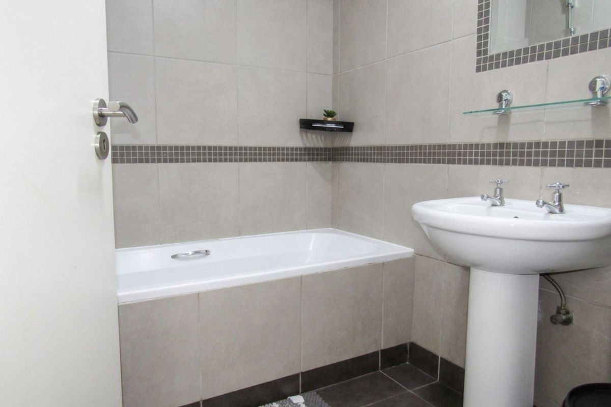 A clean, neutral-toned bathroom featuring a built-in bathtub, pedestal basin, and tiled finishes throughout.