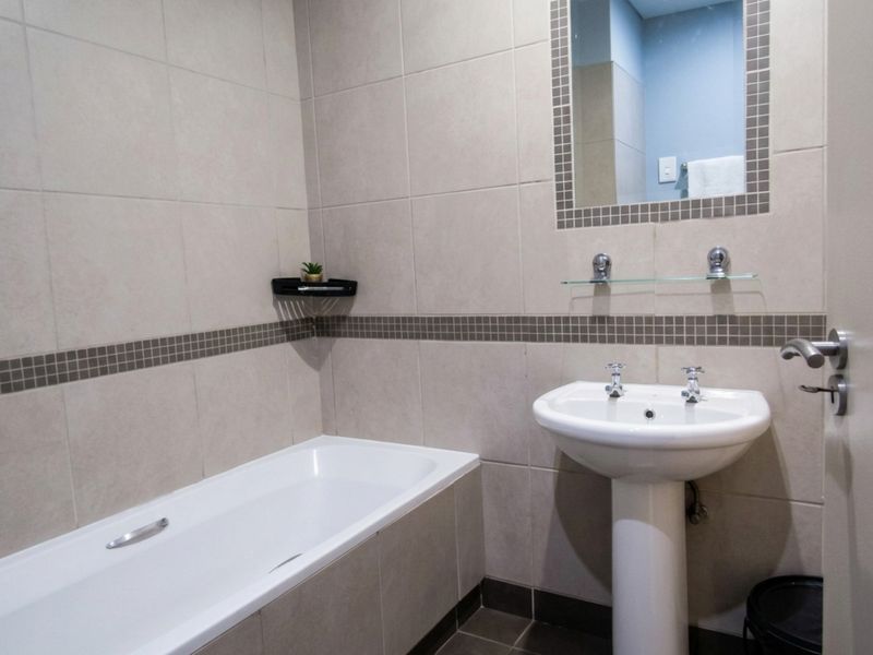 A clean, functional bathroom featuring a white pedestal basin, a standard bathtub, and neutral ceramic wall tiling with a decorative mosaic border.