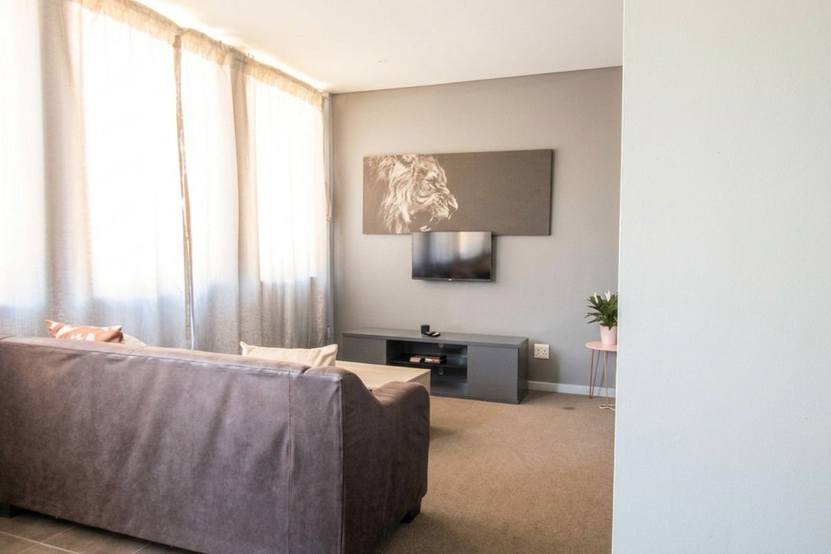 A comfortable living room featuring carpeted flooring, a large window with curtains, and a wall-mounted television setup.
