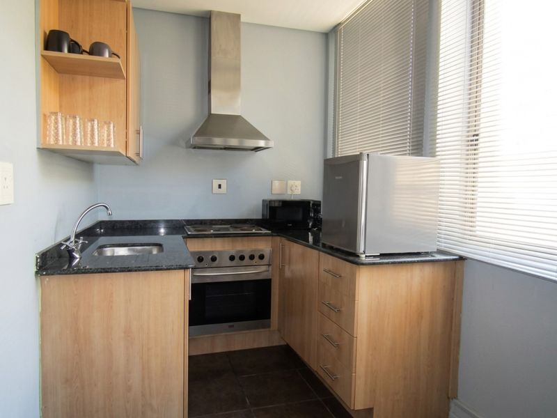Compact kitchen featuring light wood cabinetry, granite countertops, stainless steel appliances including an oven, hob, and extractor fan, with natural light from large windows.