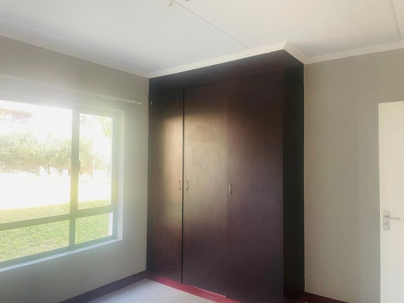 A bright, empty bedroom featuring dark wood built-in cupboards, neutral wall finishes, and a large window overlooking the garden area.
