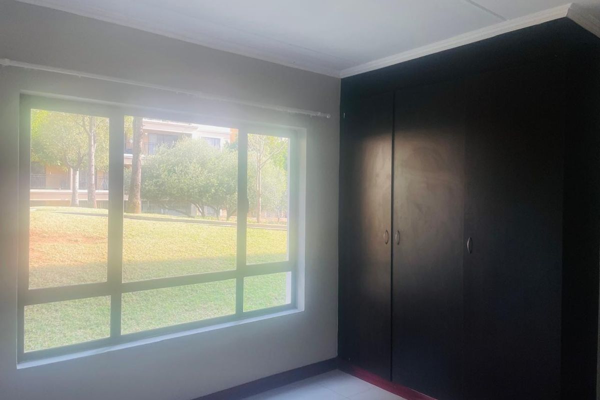 A bright, empty bedroom featuring tiled flooring, a large window with garden views, and dark built-in cupboards.