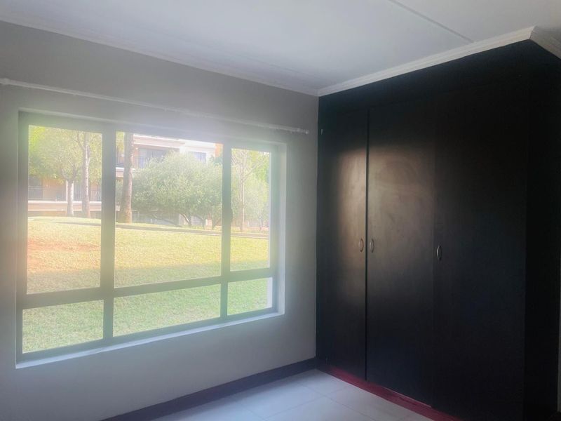 A bright, empty bedroom featuring tiled flooring, a large window with garden views, and dark built-in cupboards.