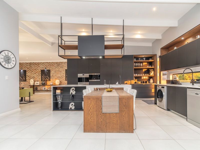 Modern open-plan kitchen featuring sleek dark cabinetry, a central wooden island, integrated appliances, and exposed ceiling beams, flowing into a living area with brick accents.