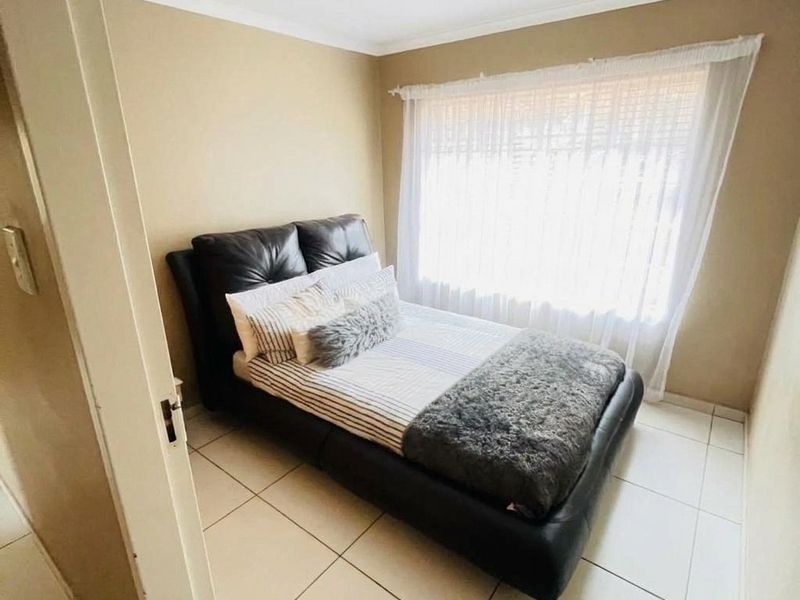 A compact bedroom featuring neutral wall tones, tiled flooring, and a large window with sheer curtains, providing a functional and well-lit sleeping space.