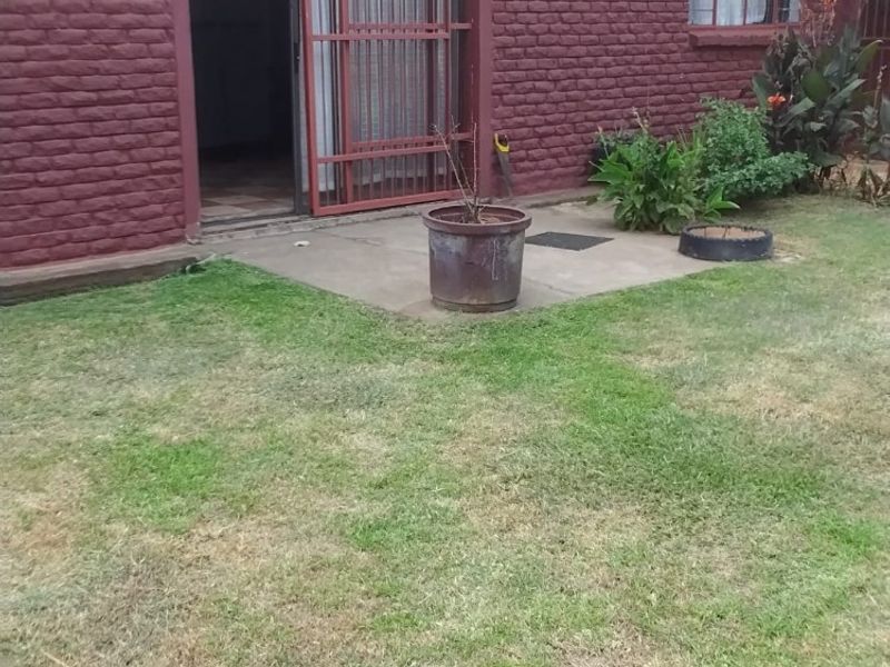 Rear view of a brick farmhouse featuring a concrete patio, security gate, and tiled roof, set against a lawn area.