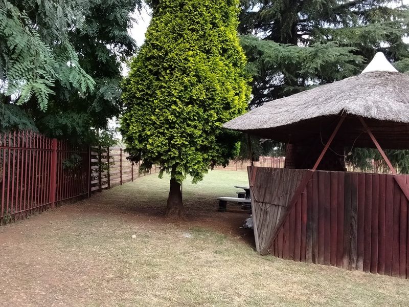 A spacious garden area featuring a traditional thatched lapa, enclosed by steel palisade and timber boundary fencing with mature trees.