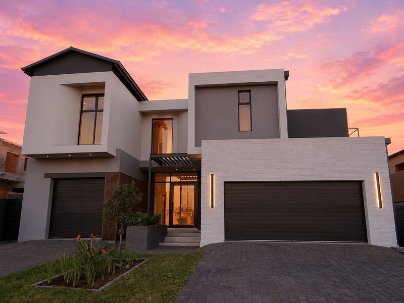 A striking modern double-story home featuring a contemporary facade, double garage, paved driveway, and elegant architectural lighting at dusk.