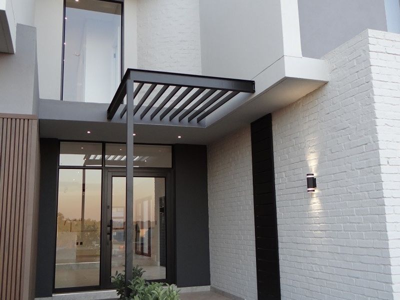 Modern front entrance featuring a painted brick facade, metal pergola, glass door, and integrated concrete planter with tiled steps.