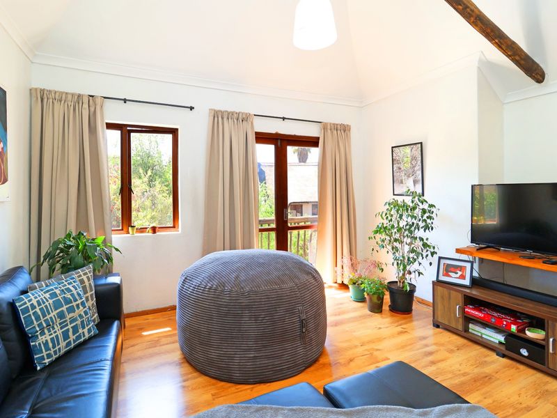 A bright living room featuring vaulted ceilings, exposed timber beams, warm timber flooring, and direct access to an outdoor balcony through timber-framed glass doors.