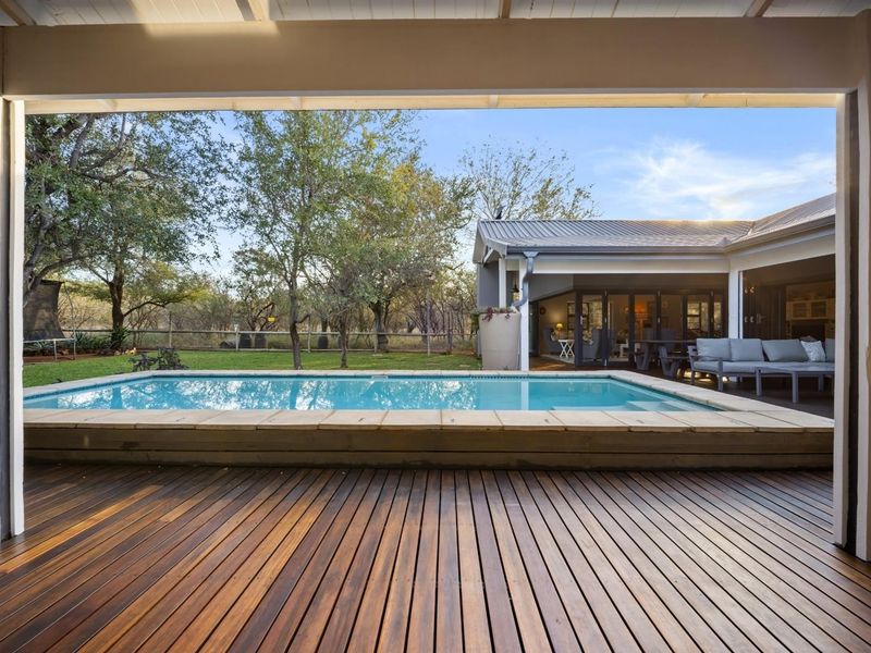 A spacious covered timber deck overlooking a swimming pool and landscaped garden, featuring seamless indoor-outdoor flow through large sliding glass doors.