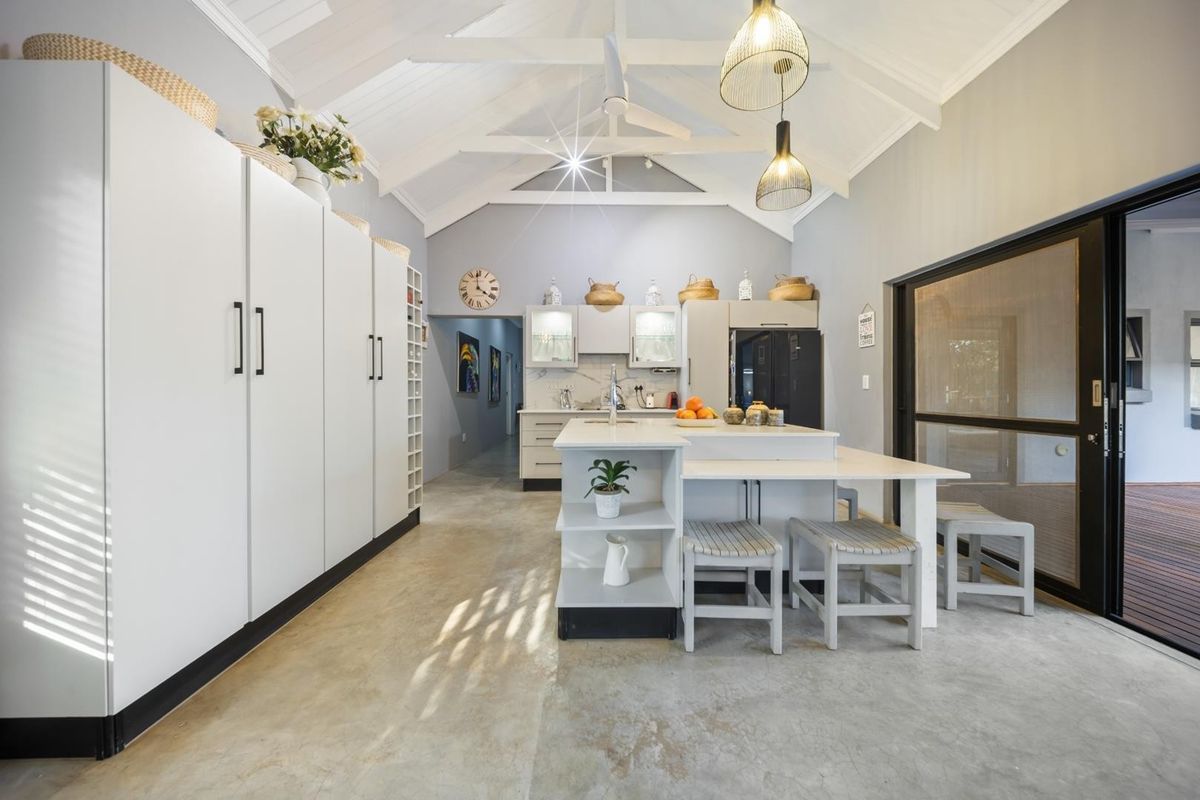 Modern kitchen featuring a central island, vaulted ceilings with exposed rafters, sleek white cabinetry, and polished concrete floors, opening onto an outdoor patio area.
