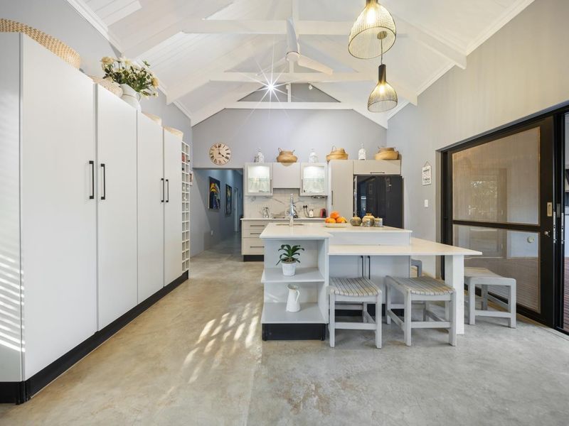 Modern kitchen featuring a central island, vaulted ceilings with exposed rafters, sleek white cabinetry, and polished concrete floors, opening onto an outdoor patio area.