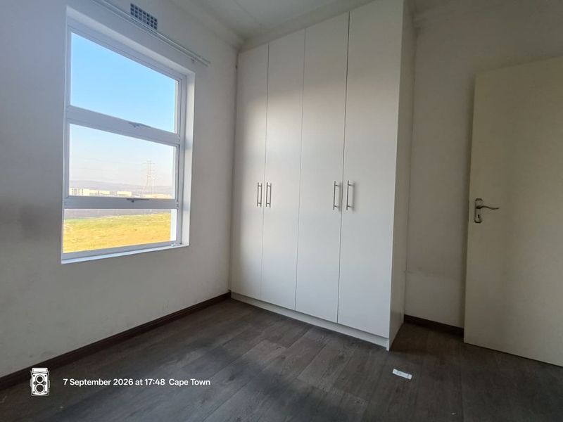A bright, empty bedroom featuring modern laminate flooring, built-in white cupboards, and a large window with natural light.