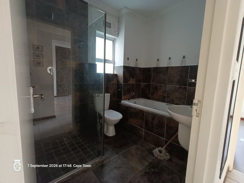 A functional bathroom featuring a glass shower enclosure, built-in bathtub, pedestal basin, and toilet with dark tiled finishes throughout.