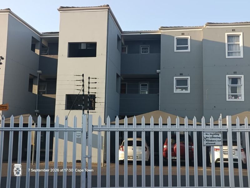 Front elevation of a multi-story apartment complex featuring a secure gated entrance, covered parking bays, and modern grey facade finishes.