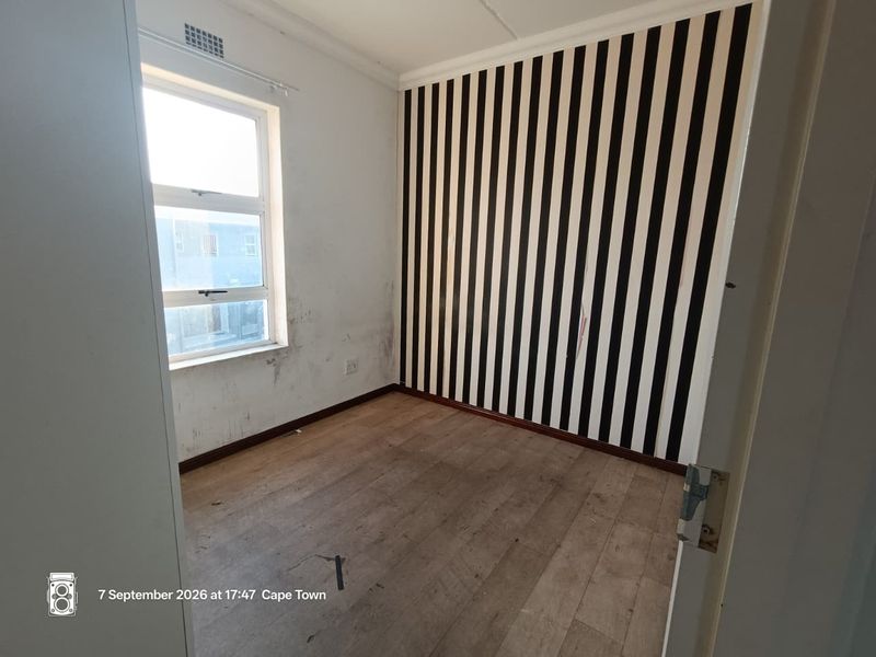An empty bedroom featuring laminate flooring and a window, currently showing signs of wall staining and damaged wallpaper requiring cosmetic attention.