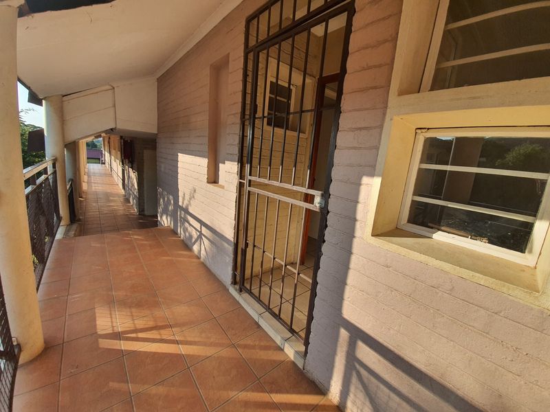 A covered exterior walkway or balcony featuring tiled flooring, brick walls, and a metal security gate entrance.
