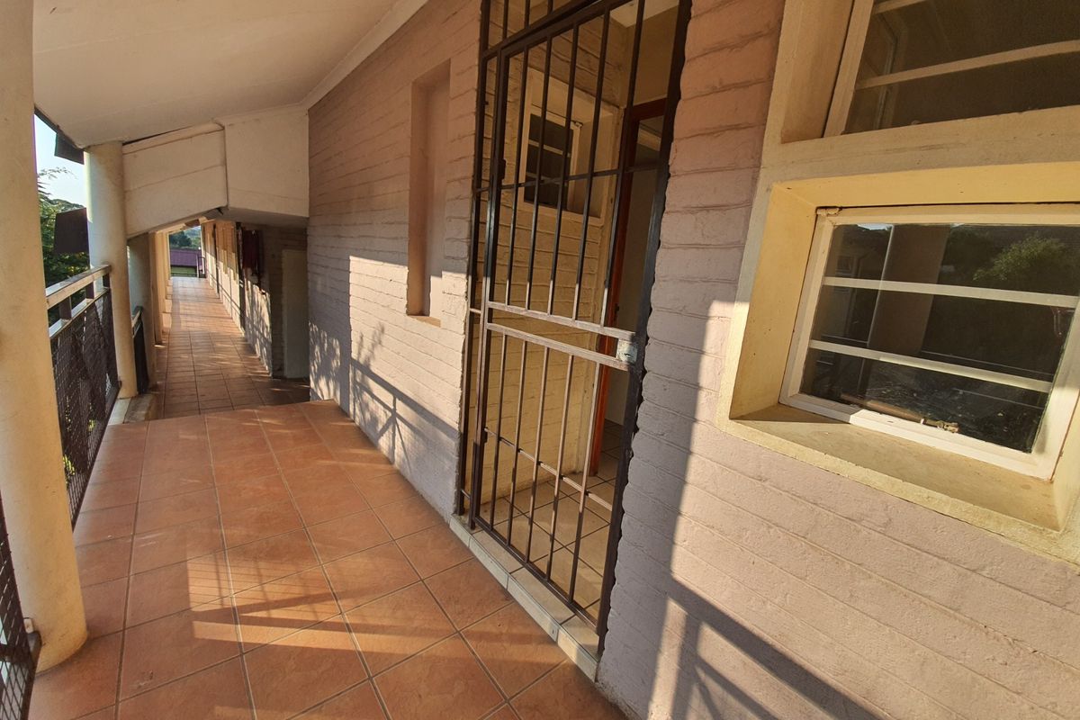 A covered exterior walkway or balcony featuring tiled flooring, brick walls, and a metal security gate entrance.