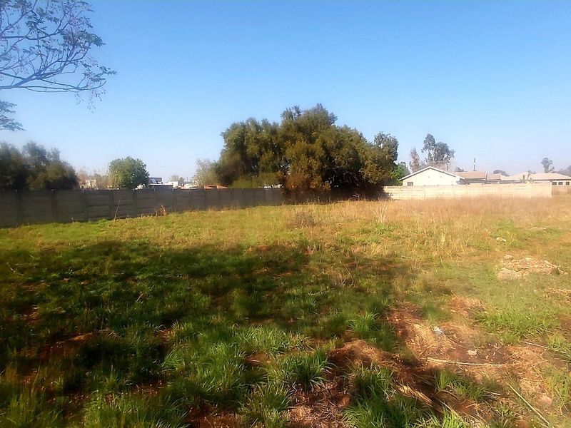 A vacant residential plot featuring natural grass coverage, mature trees, and a precast concrete boundary wall along the perimeter.
