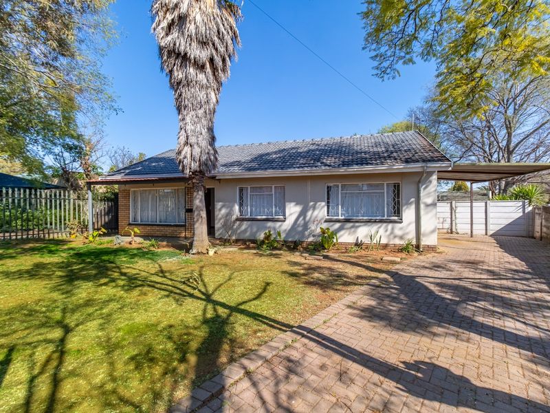 Front view of a single-story house featuring a paved driveway, carport, tiled roof, and established garden with security fencing.
