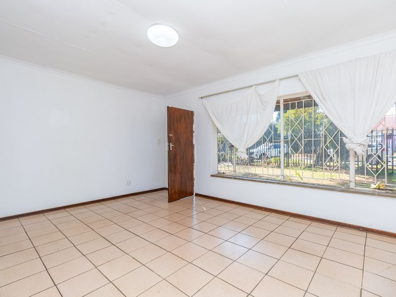 A bright, empty living room featuring neutral tiled flooring, large windows with security bars, and a simple ceiling light fixture.