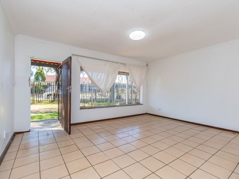 An empty, sunlit living room featuring neutral tiled flooring, a security gate entrance, and burglar bars on the window.