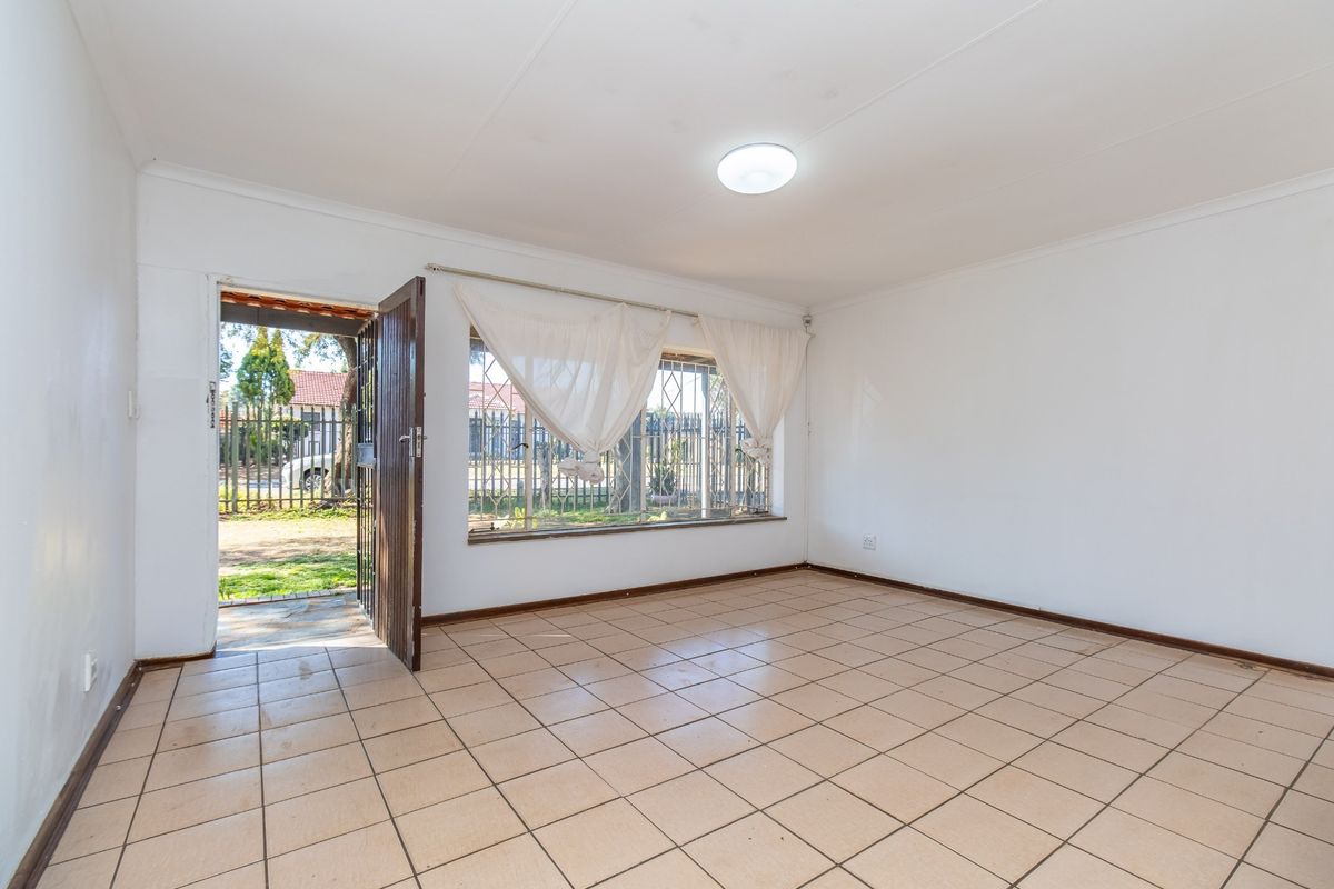 An empty, sunlit living room featuring neutral tiled flooring, a security gate entrance, and burglar bars on the window.