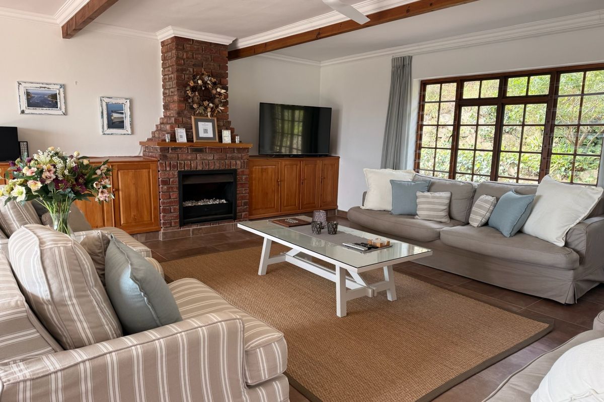A cozy living room featuring exposed timber ceiling beams, a classic brick fireplace, built-in wooden cabinetry, and large lattice-style windows overlooking the garden.