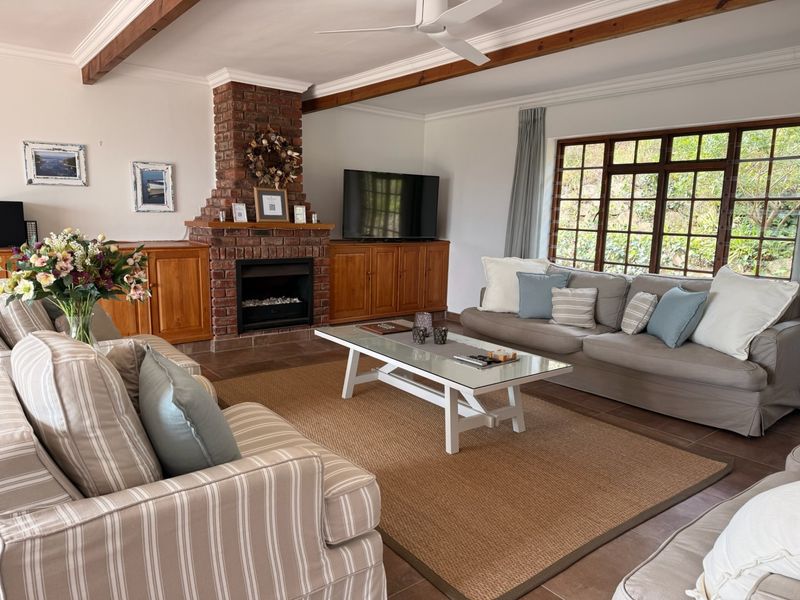 A cozy living room featuring exposed timber ceiling beams, a classic brick fireplace, built-in wooden cabinetry, and large lattice-style windows overlooking the garden.