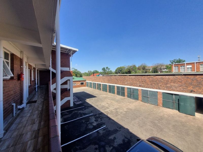 An exterior view of the apartment complex rear area, featuring a tiled walkway, paved parking bays, and secure metal storage or parking enclosures.