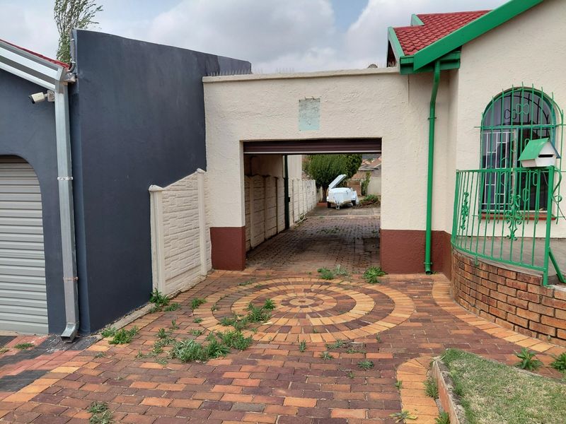 Front exterior view showing a paved driveway, boundary wall with security spikes, and access to a covered parking area.