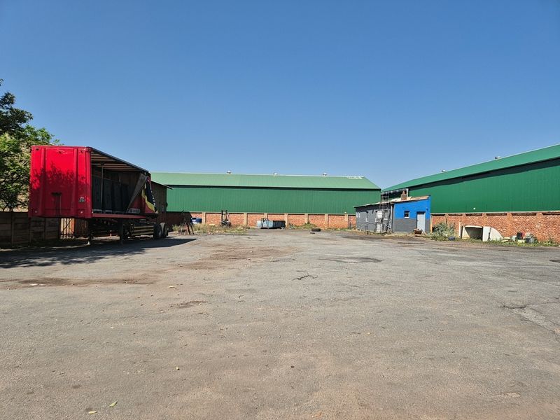 Spacious industrial yard featuring asphalt surfacing, brick perimeter walls, and large warehouse structures with green metal roofing.
