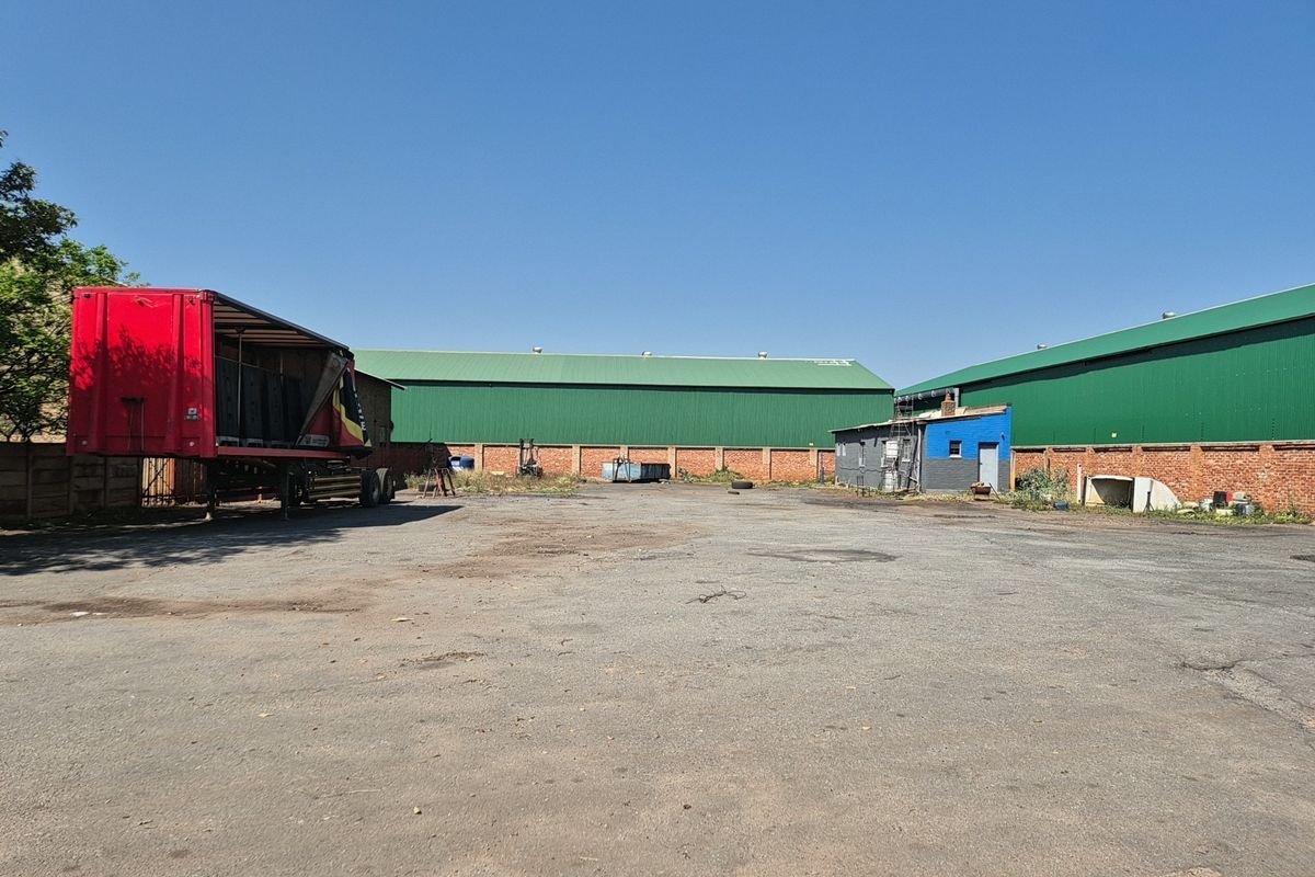 Spacious industrial yard featuring asphalt surfacing, brick perimeter walls, and large warehouse structures with green metal roofing.