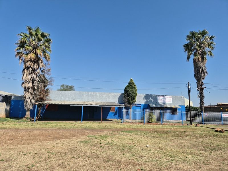 Front elevation of an industrial property featuring a painted brick facade, metal roof, covered entrance area, and perimeter steel security fencing.