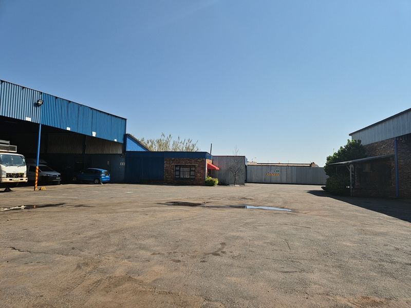 Spacious industrial yard featuring corrugated metal warehouse structures, brick facade sections, and ample open-air parking space.